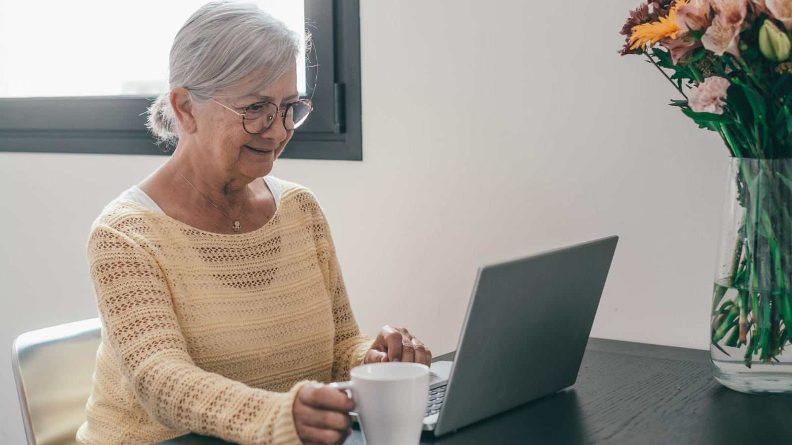 Elderly woman sitting at a table holding a mug in one hand whilst her other hand is resting on a laptop in front of her