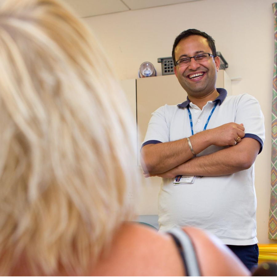 image of a male clinician standing smiling with his arms folded in front of a female patient who is facing him. 