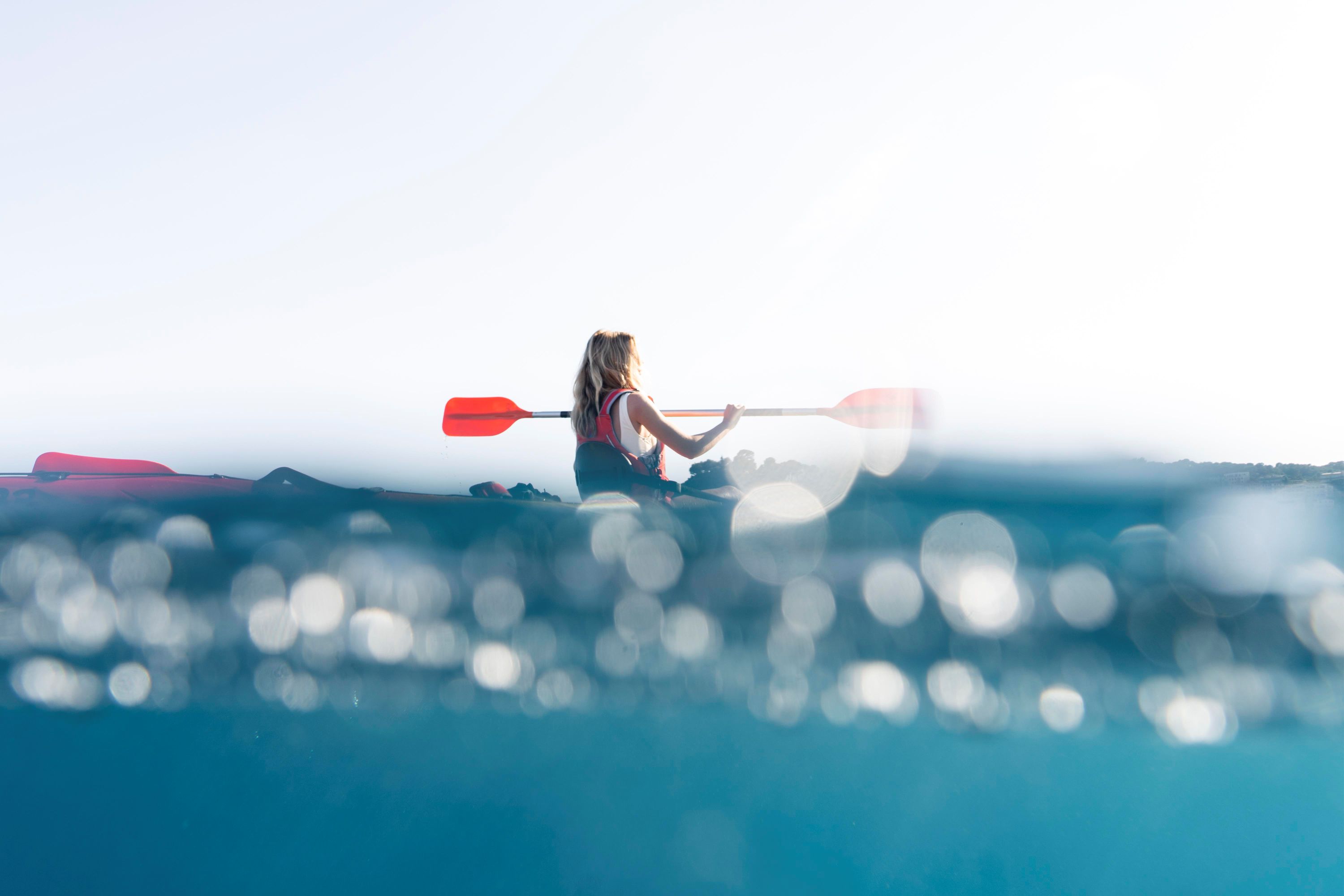 A young blonde woman in a life jacket is sitting in a canoe with the sea rippling around her. she holds orange ores and is paddling with them. 