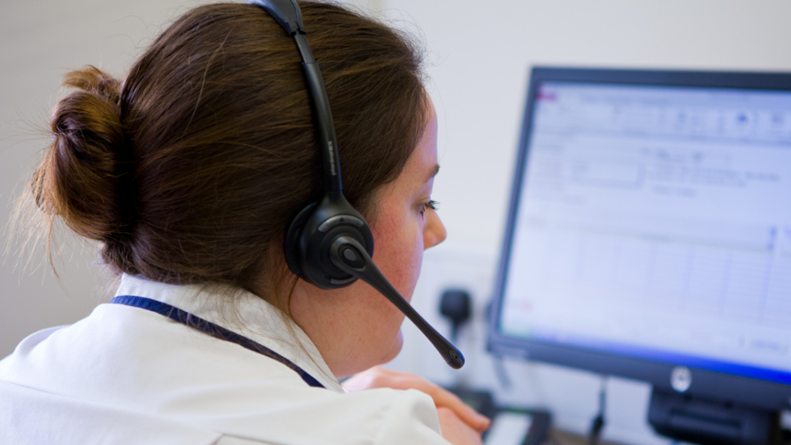 Woman in a white shirt with a headset on, faces away from the camera and towards a computer screen. She is clasping her hands together in front of her.